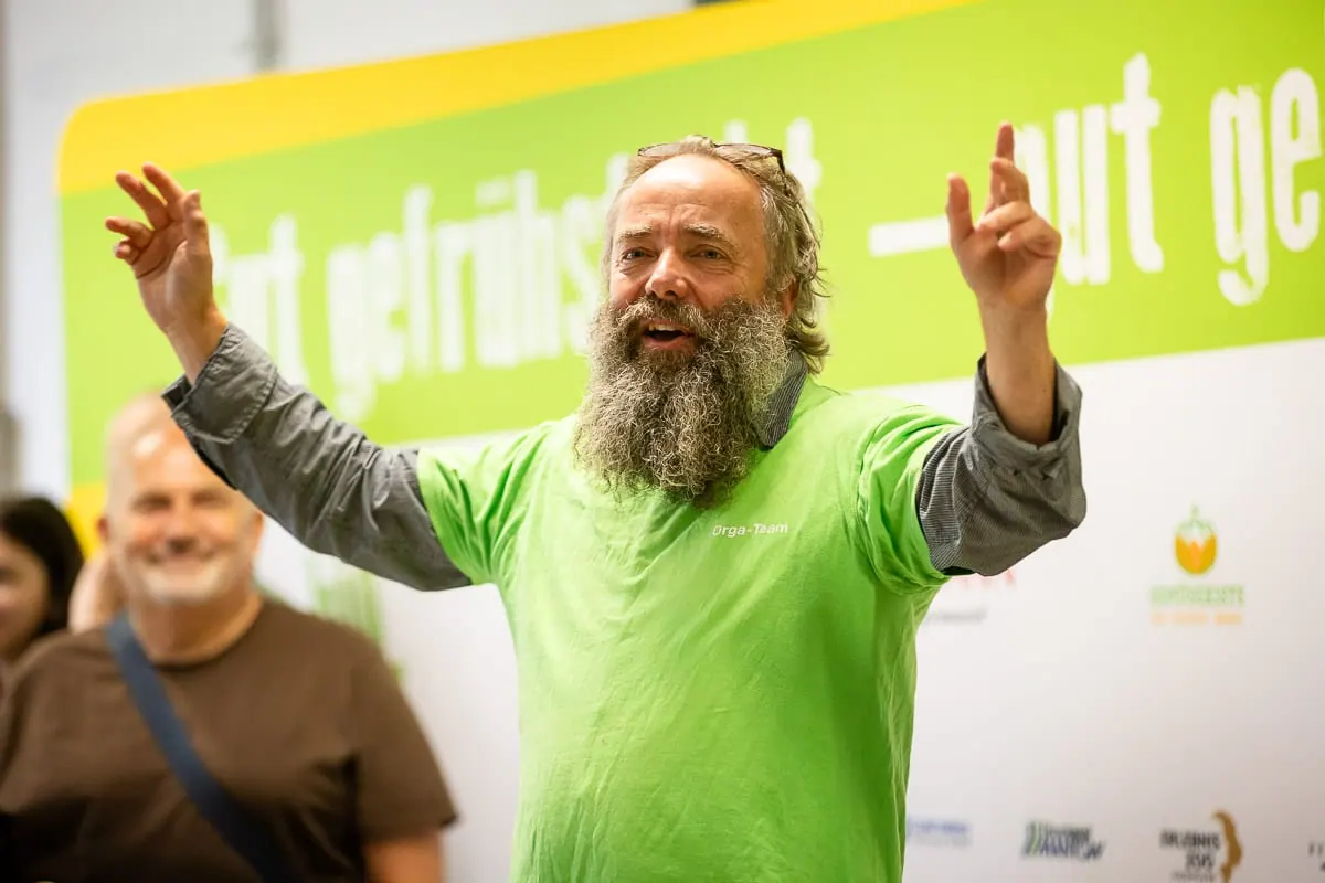 Mann in grüner Jacke mit erhobenen Händen vor einer Bildwand